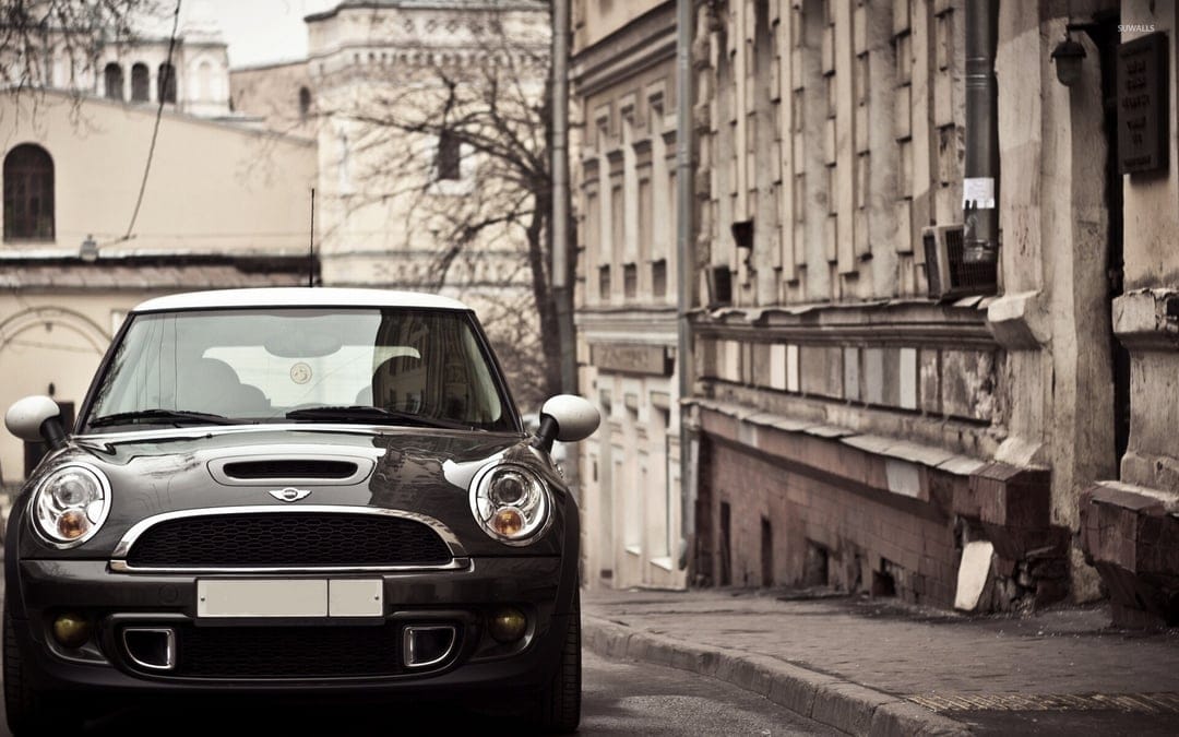 Front view of a dark Mini Cooper with a white roof parked on a paved street next to a textured stone building.