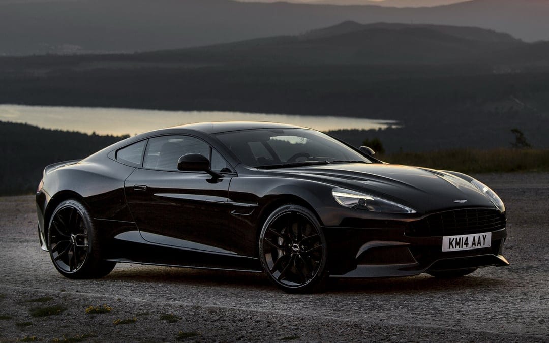 Black Aston Martin Vanquish parked on gravel with mountains and a lake in the background at sunset.