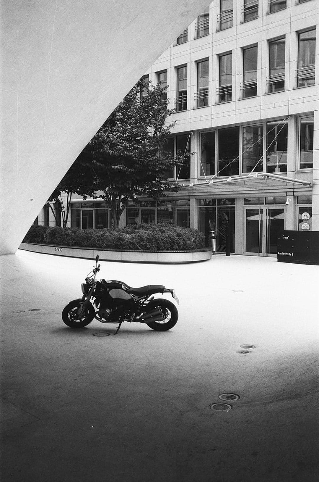 Black motorcycle parked on a paved courtyard in front of a modern glass building with curved concrete features.