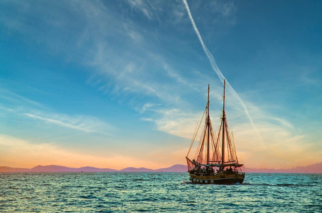Two-masted sailboat on calm orange water during sunset with wispy clouds and a distant island silhouette.