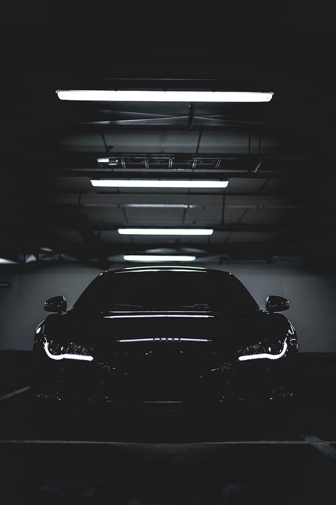 Black Audi luxury car with glowing headlights parked under fluorescent lights in a dark underground garage.