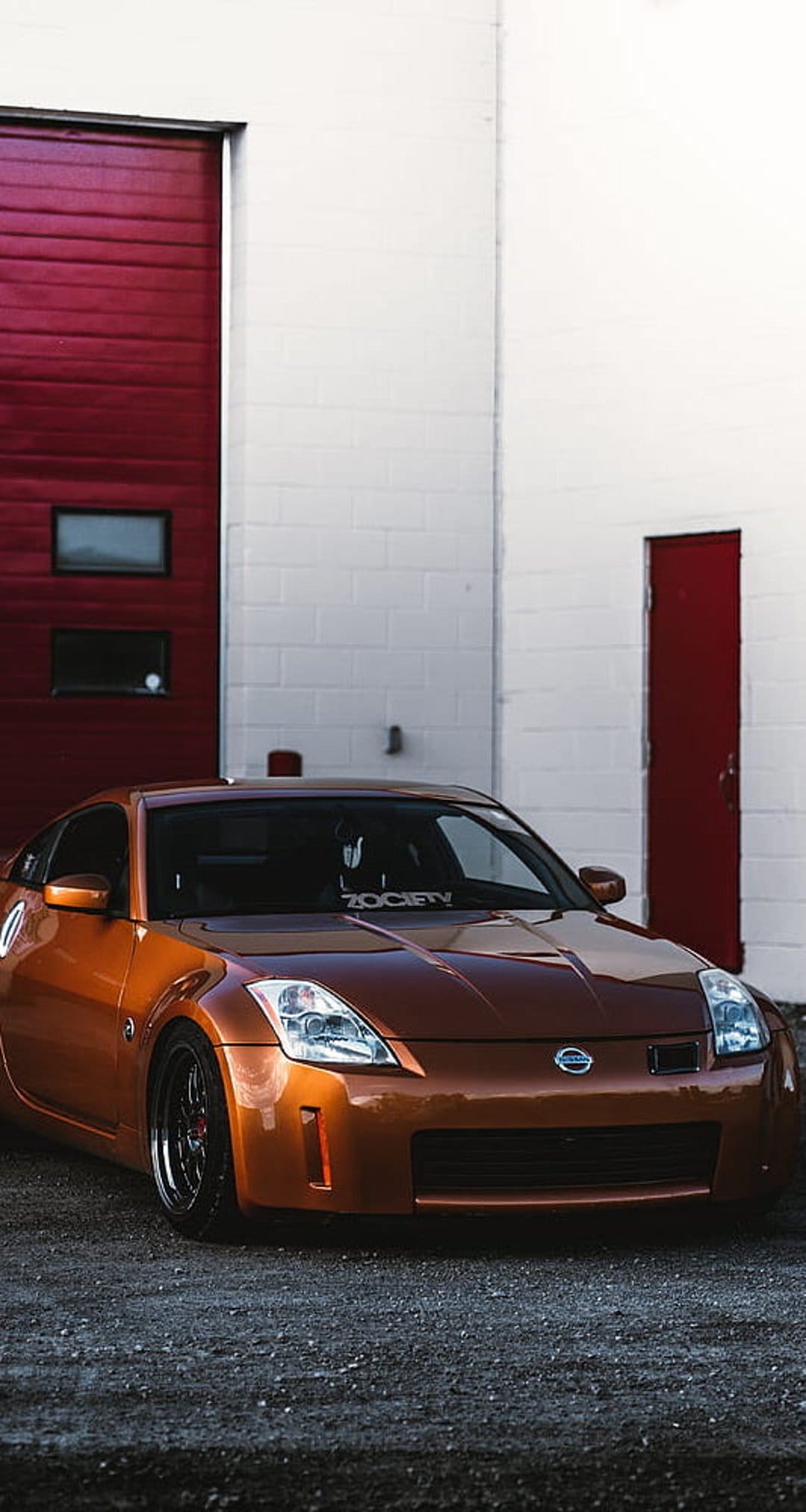 Nissan 350Z in metallic orange parked on asphalt in front of large red doors at an industrial facility.