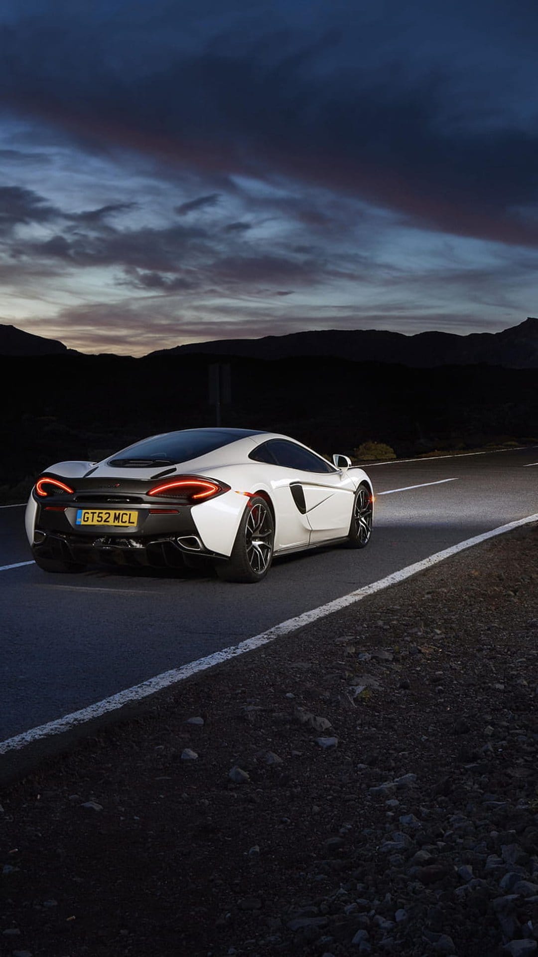 White McLaren supercar rear view with glowing red taillights on a winding desert road under a twilight sky.