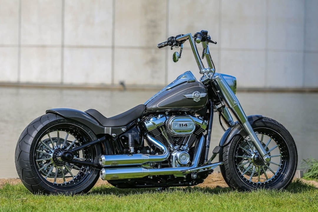 Customized gray Harley-Davidson motorcycle with chrome exhaust and ape hangers parked on green grass by water.