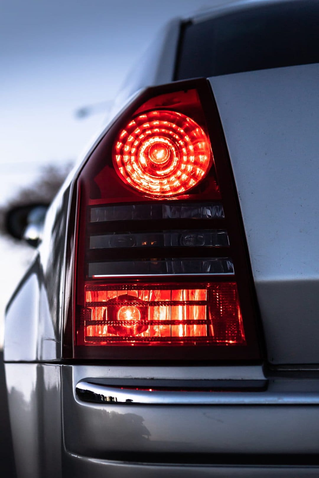 Red circular car taillight glowing at dusk with concentric light rings and reflections on a dark metal fender.