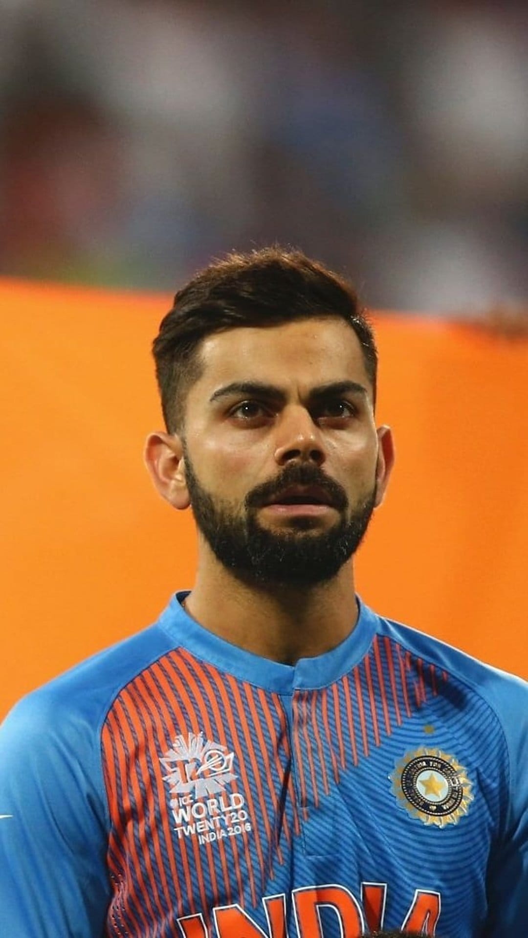Virat Kohli in a blue India cricket jersey with orange collar details stares forward with a focused expression.