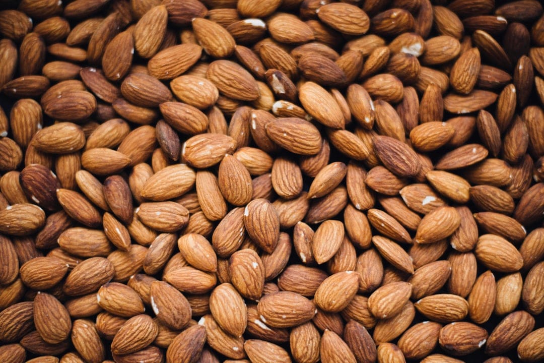 Raw almonds with brown skins are piled together in a macro overhead view showing varied oval shapes and textures.