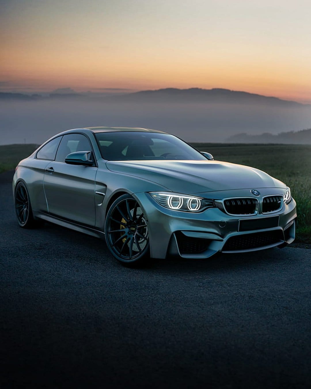 Silver BMW M4 Coupe parked on a paved road during a golden sunrise with misty mountains in the background.