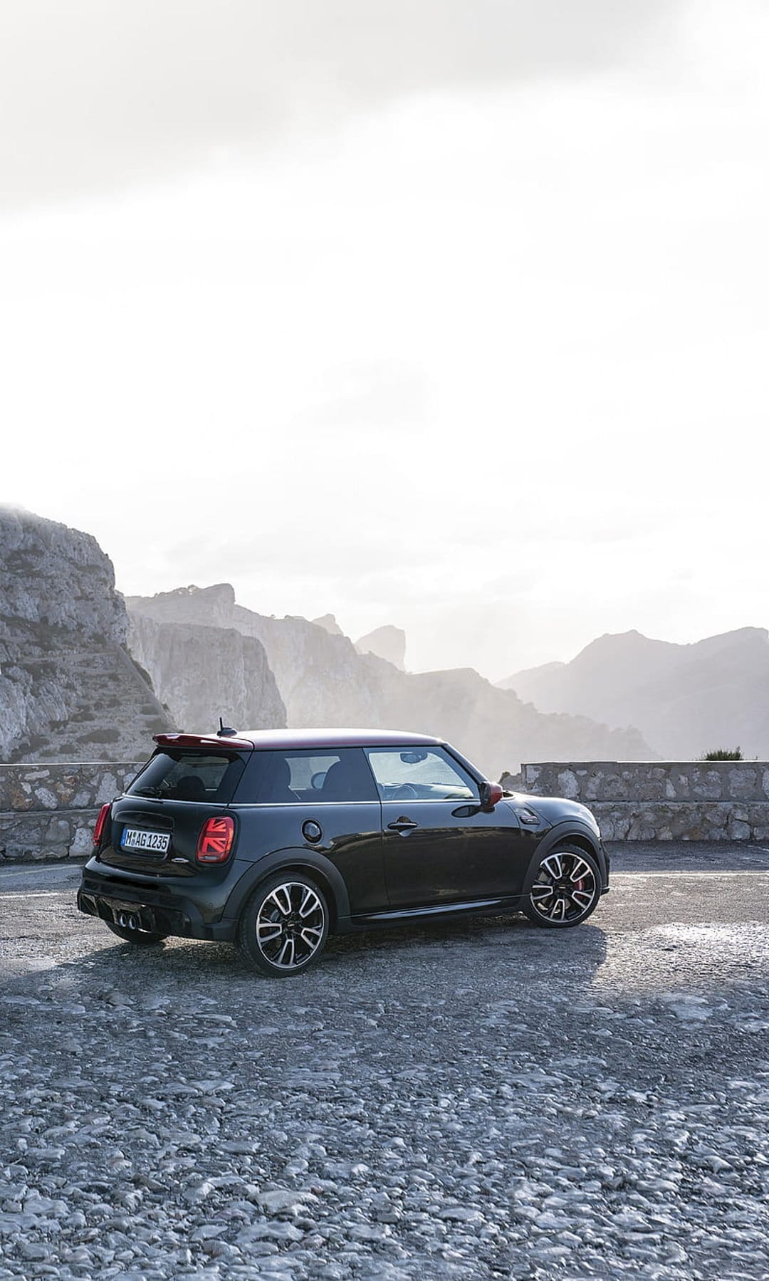 Dark Mini Cooper parked on a gravelly mountain overlook with foggy peaks and a dramatic, overcast sky.
