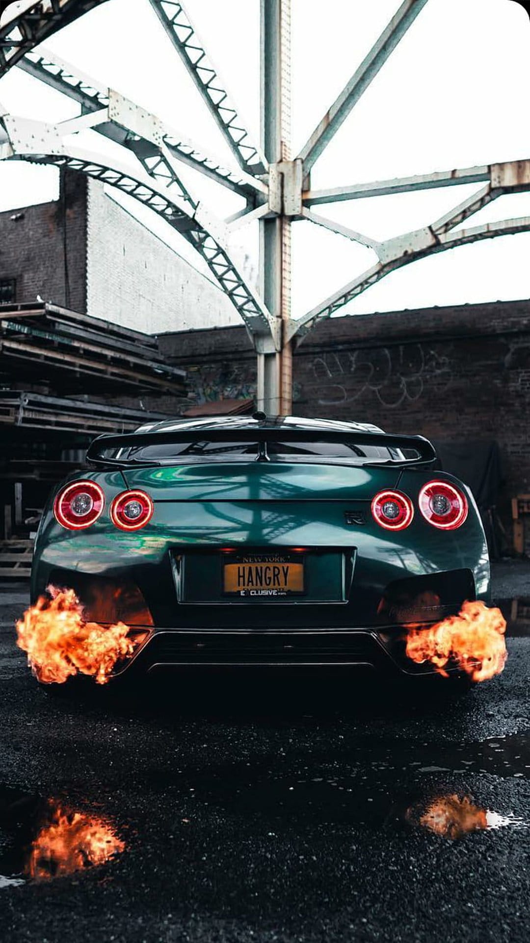 Nissan GT-R with orange exhaust flames on wet asphalt under a steel bridge at night with hangry license plate.