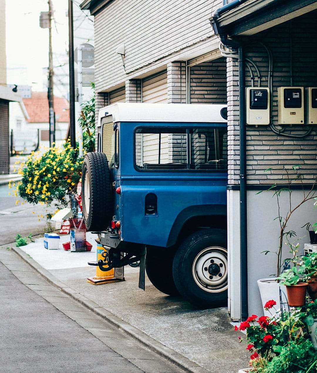 Blue Land Rover Defender with a white roof parked near a grey brick house and green bushes with red flowers.