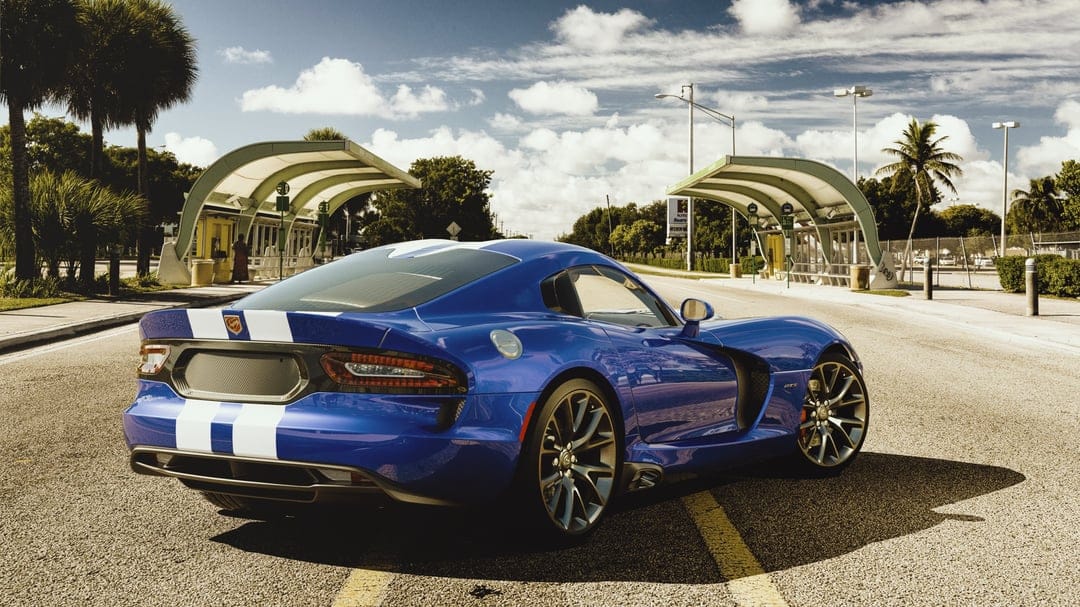 Blue sports car with white racing stripes parked on a sunlit road next to palm trees and modern bus shelters.