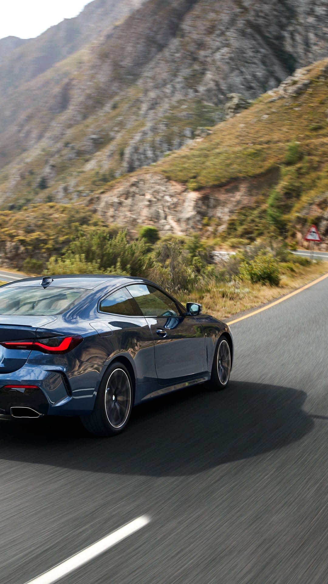 Dark blue sport coupe driving on a winding mountain road with rugged peaks and motion-blurred asphalt.