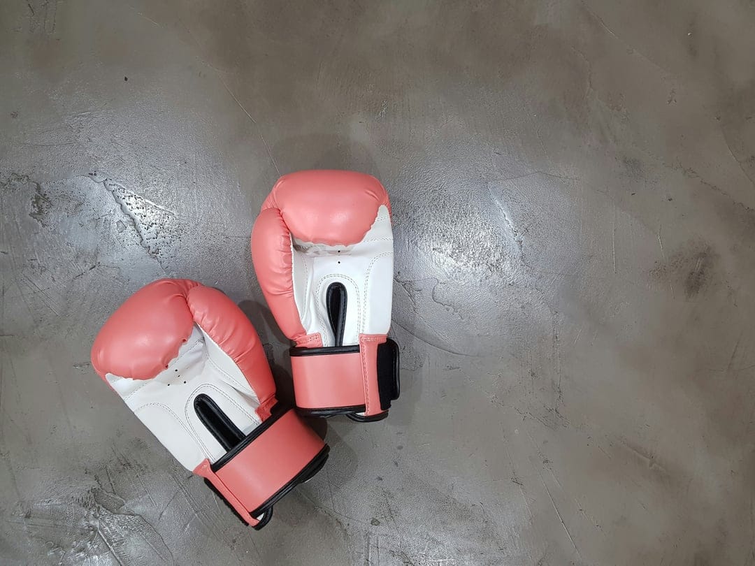 Pink and white boxing gloves positioned diagonally on a gray textured concrete floor with soft shadows.