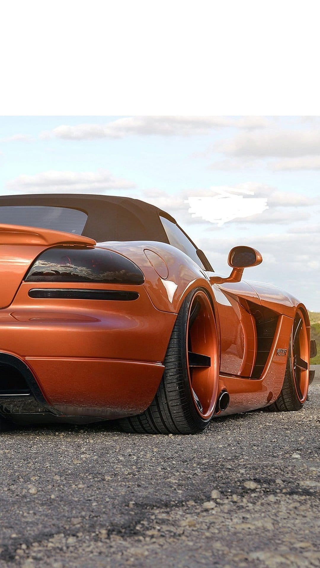 Orange Dodge Viper convertible with custom wheels and tinted taillights parked on a grey gravel road under clouds.