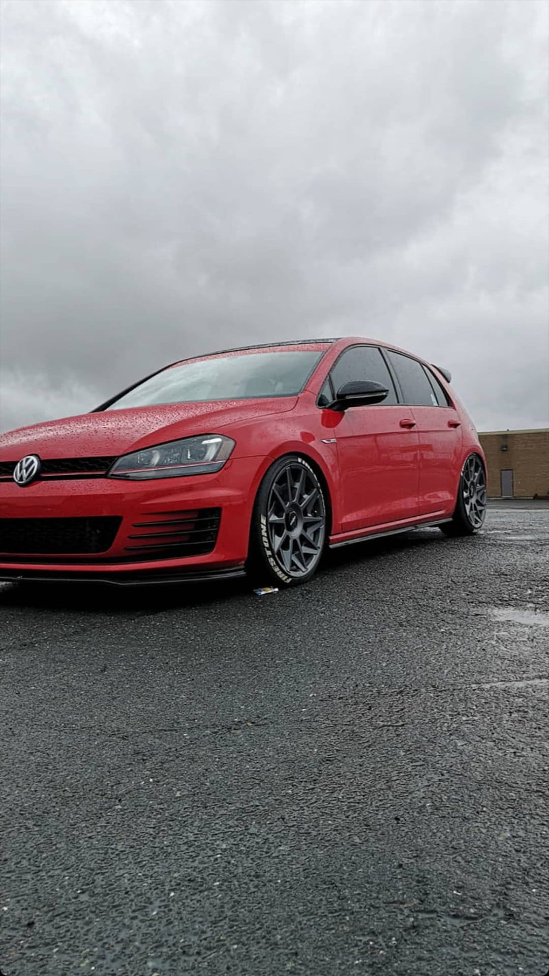 Red Volkswagen Golf GTI with dark multi-spoke wheels parked on wet asphalt under a cloudy, overcast sky.