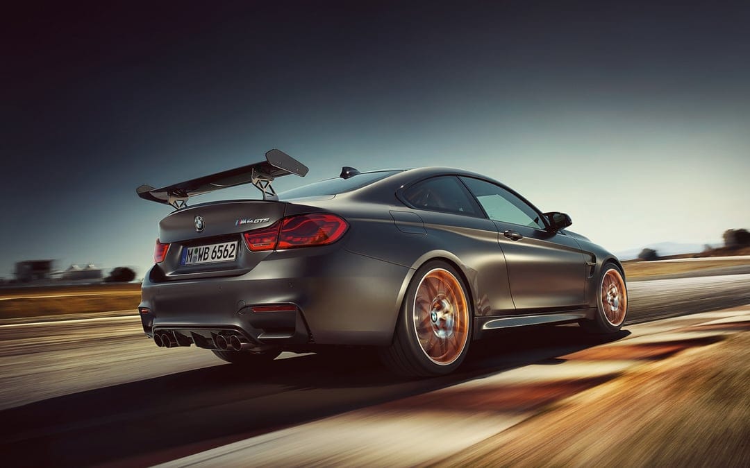 Matte grey BMW M4 GTS sports car with orange rims racing on a track with motion blur under a dramatic dark sky.