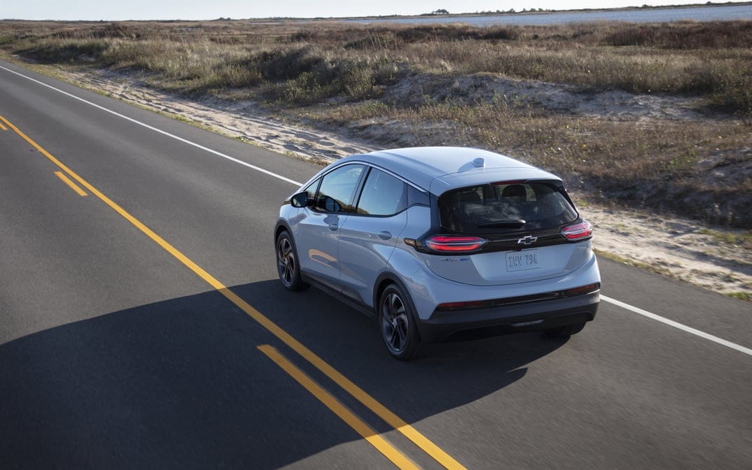 Light blue Chevrolet Bolt EV driving on a paved coastal road between sandy dunes and dry grass under bright sunlight.