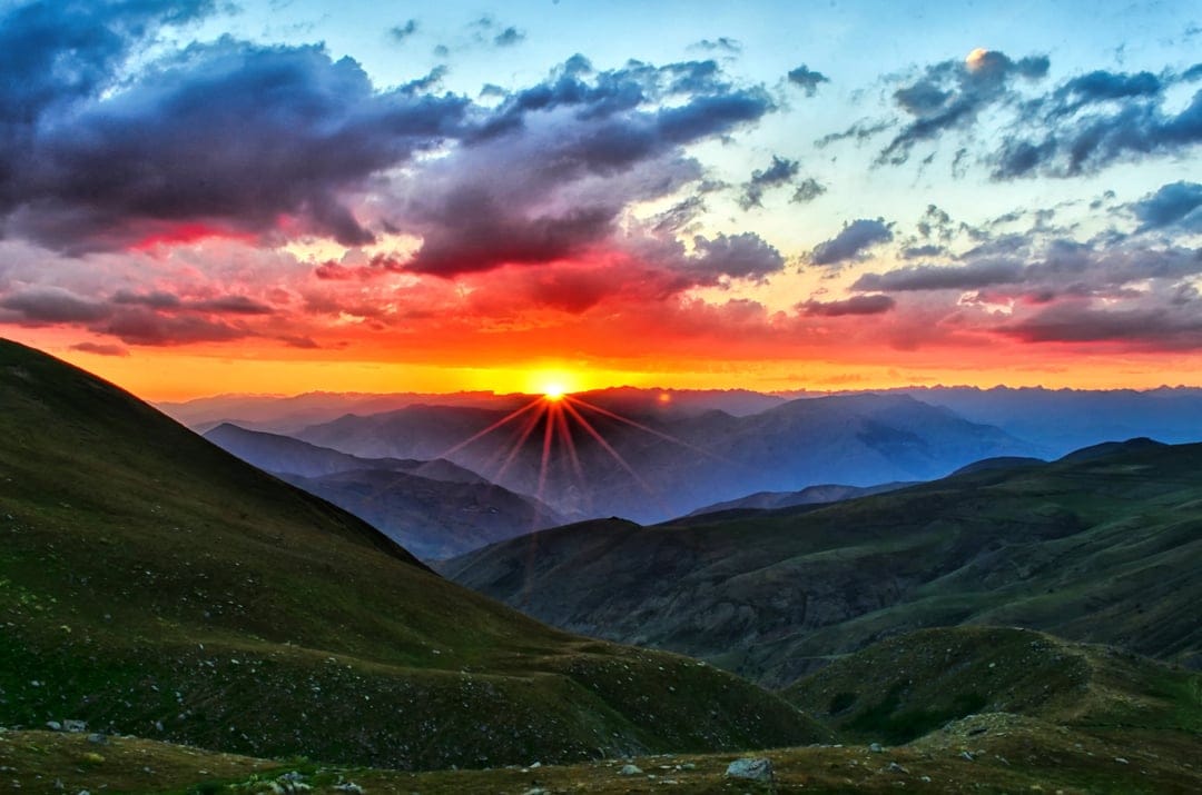 Jagged mountain peaks under a fiery orange sunset with sunbeams breaking through dense, dark purple clouds.