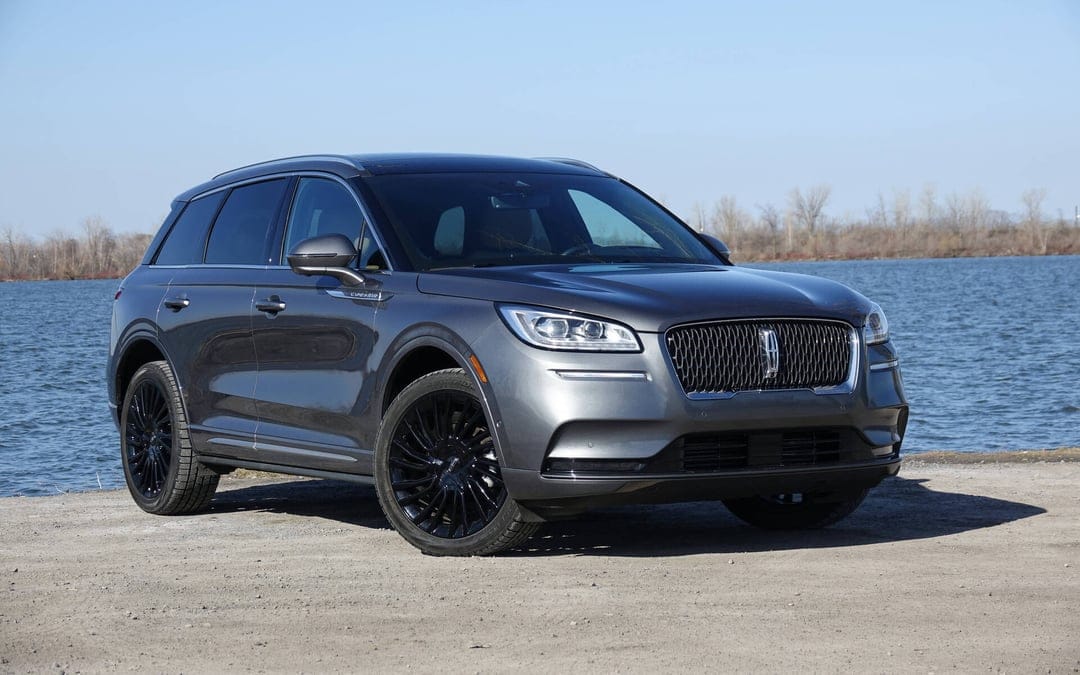 Dark gray Lincoln Corsair SUV with black rims parked on a dirt shore beside a calm blue lake under a clear sky.