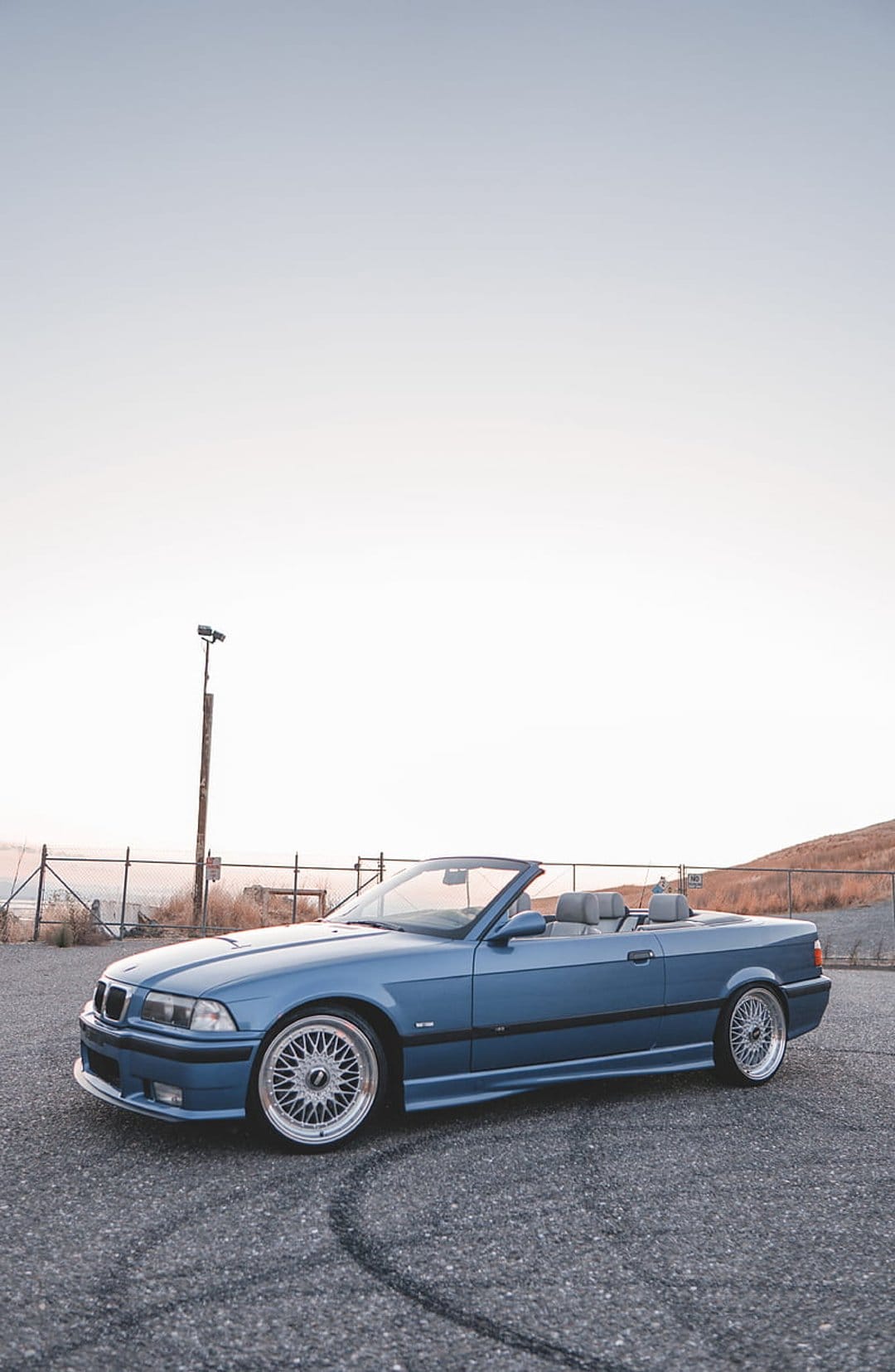 Blue BMW E36 convertible parked on rough asphalt under a pale sky during a hazy morning with visible tire marks.