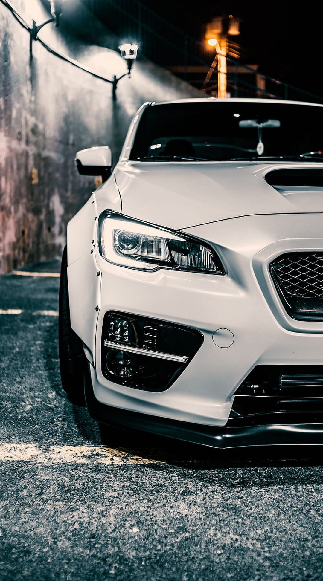 White Subaru WRX STI with glowing headlights and a widebody kit parked on a dark asphalt street at night.