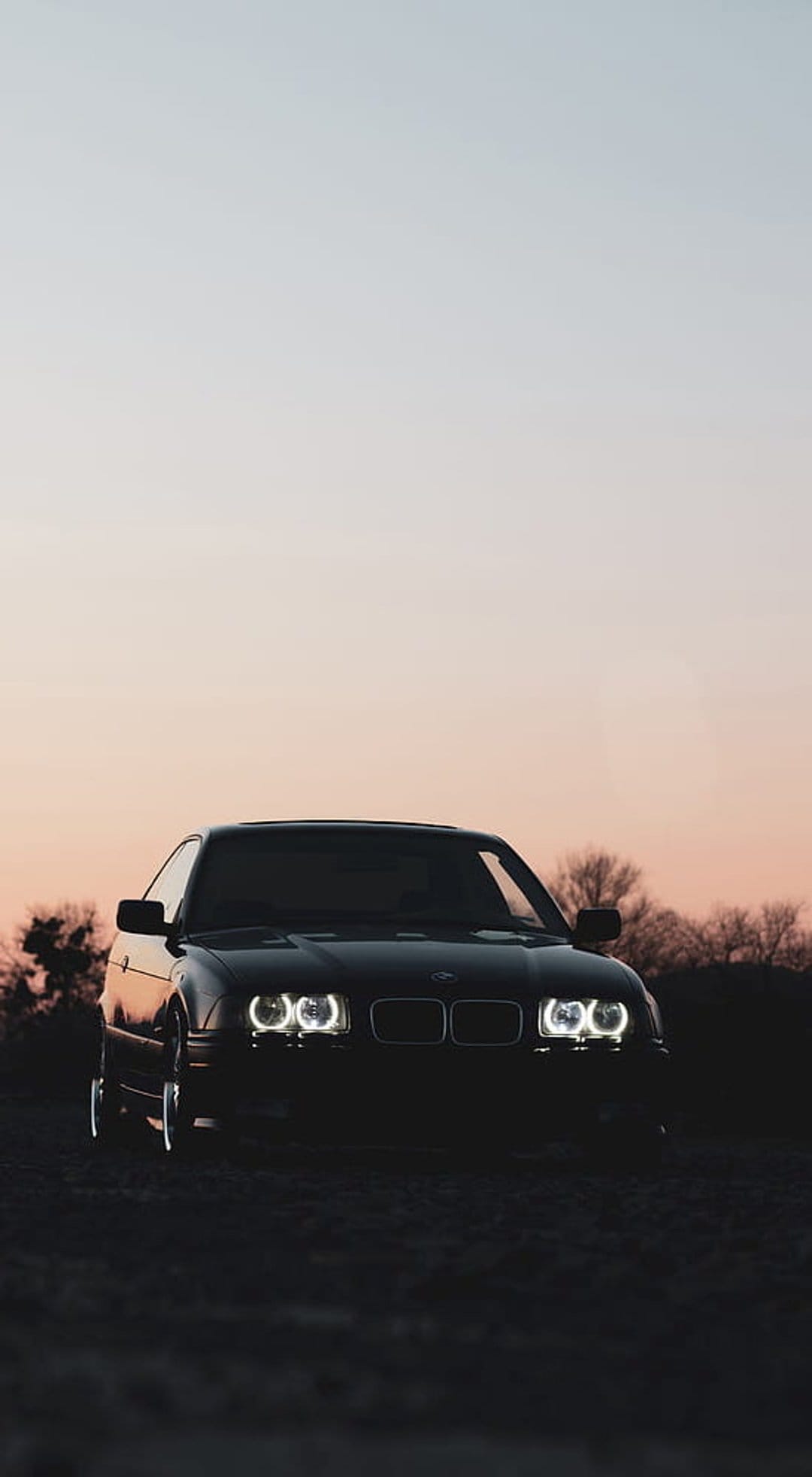 Black BMW E36 coupe with glowing halo headlights parked on rough terrain against a colorful gradient sunset sky.