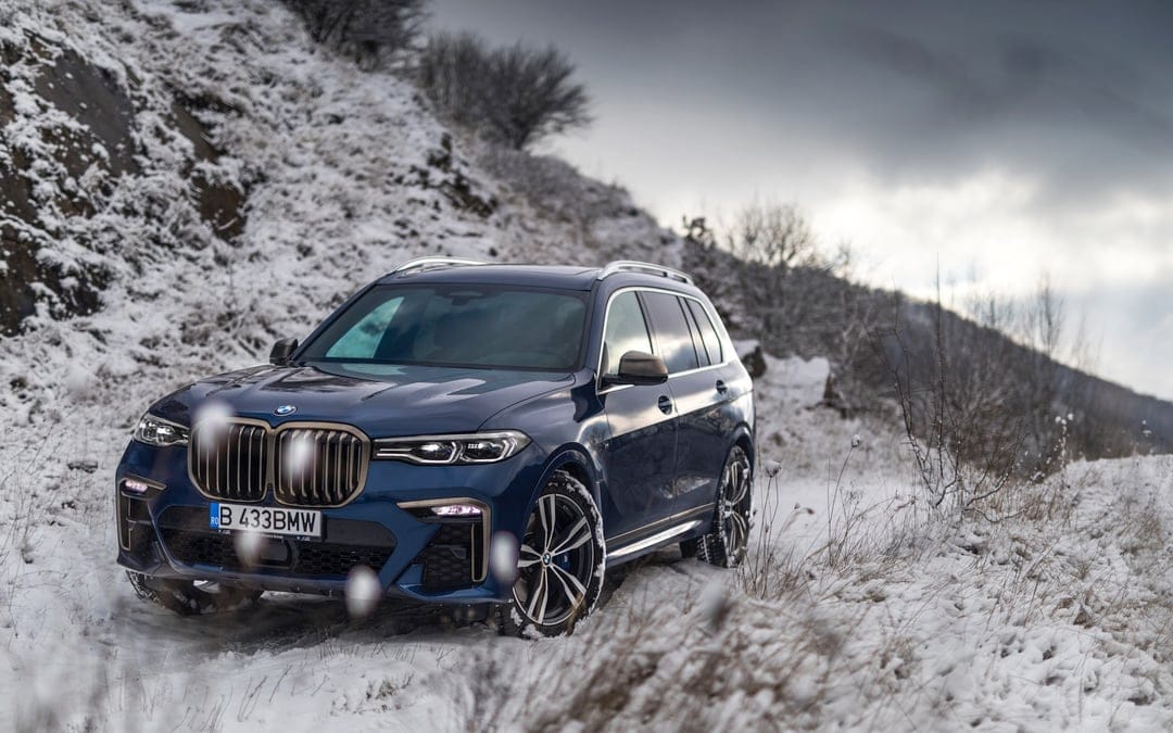 Blue BMW X7 SUV with bronze grille parked on a snowy mountain trail against a cloudy winter sky background.