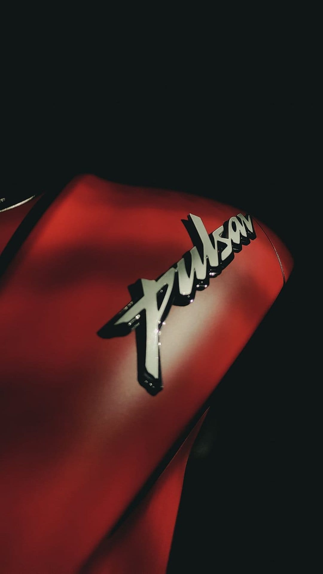 Pulsar motorcycle logo in chrome lettering mounted on a glossy red fuel tank with sharp highlights and shadows.