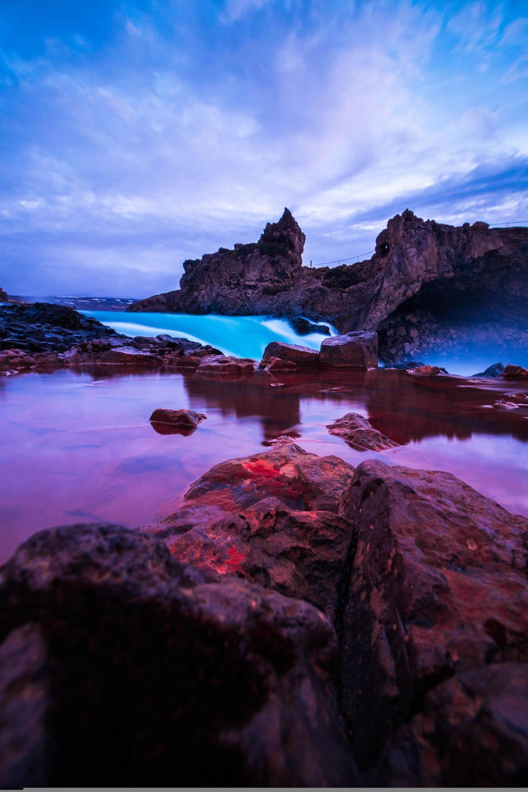 Turquoise waterfall cascading into a purple reflection pool surrounded by dark jagged rocks under a twilight sky.
