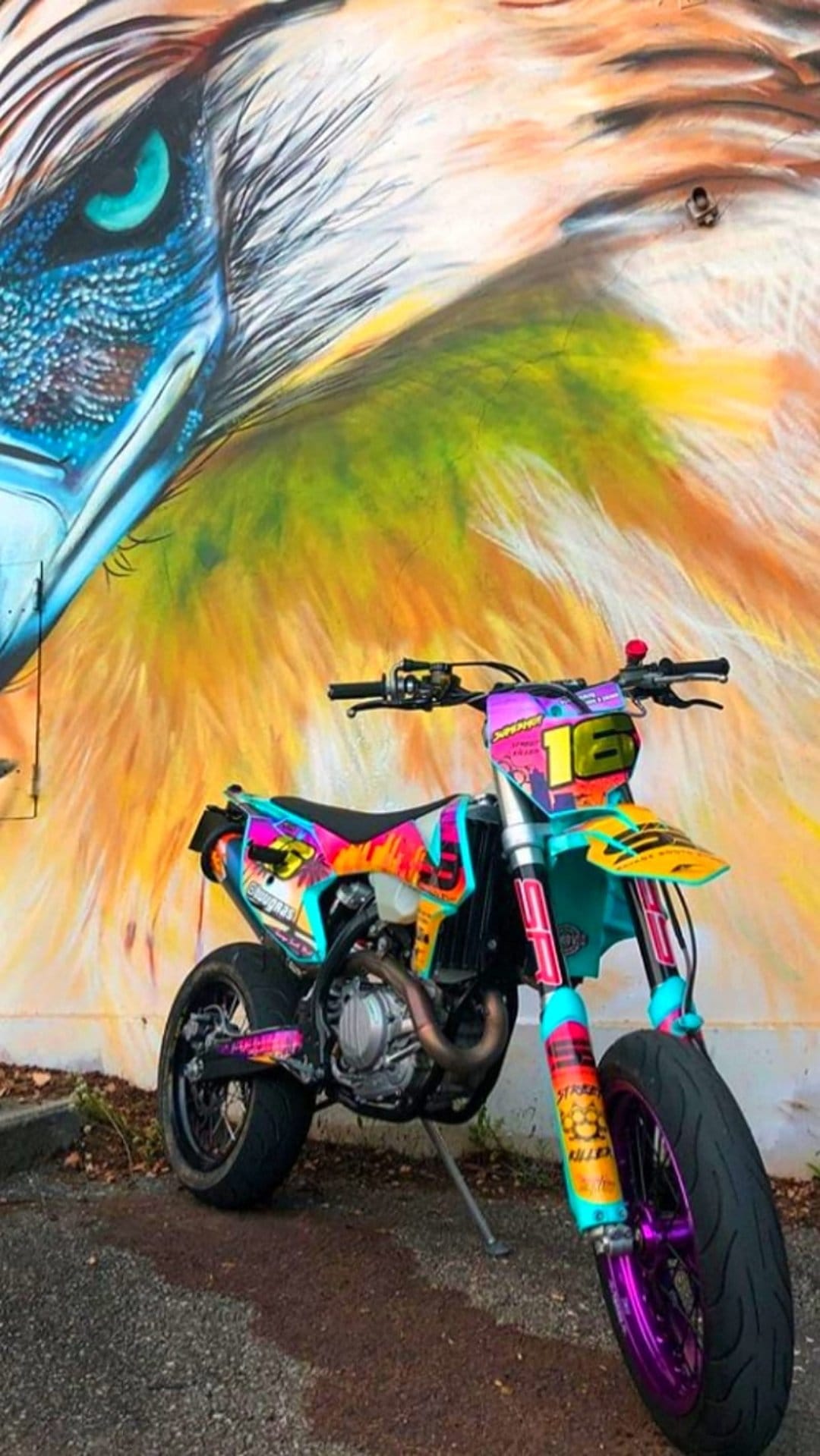 Supermoto motorcycle with a colorful wrap parked in front of a large spray-painted eagle mural on a city wall.