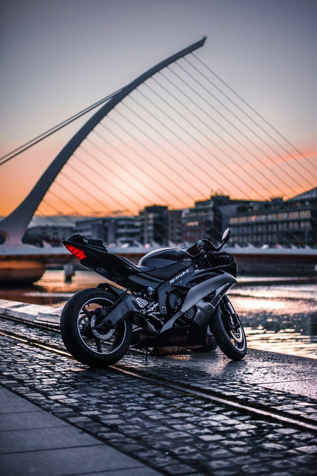 Black sportbike on cobblestones with the harp-shaped Samuel Beckett Bridge under a glowing sunset in Dublin.