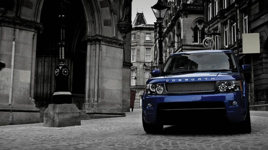 Blue Range Rover Sport with Cosworth lettering on a grayscale cobblestone street by historic stone buildings.