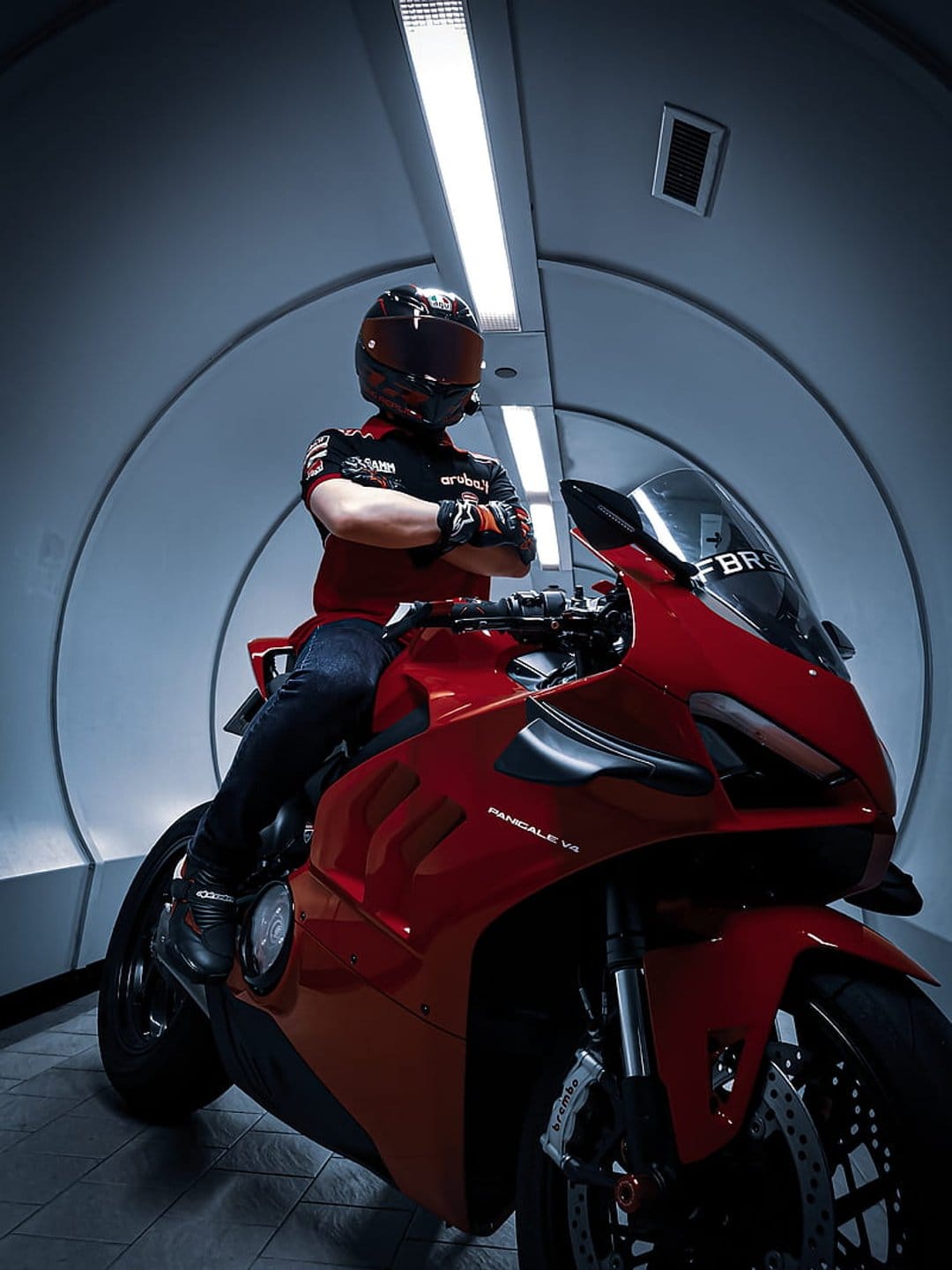 Motorcyclist in red racing gear sits on a Ducati bike inside a bright, futuristic cylindrical tunnel.