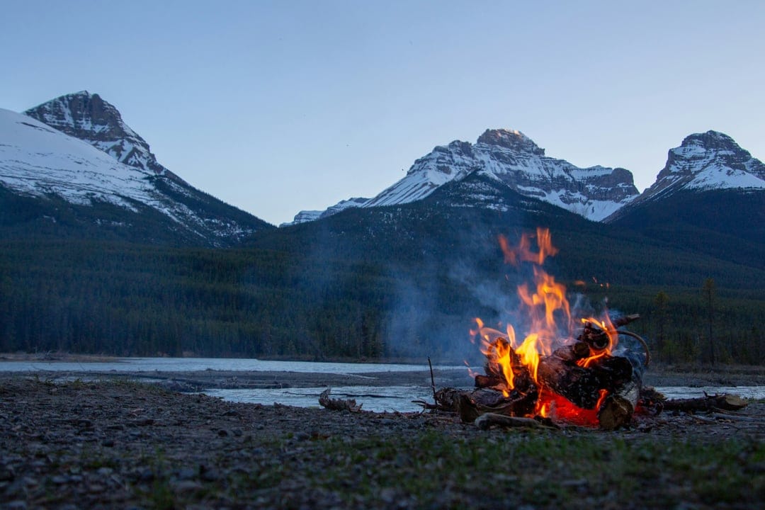 Glowing campfire on a rocky lakeshore with snow-dusted mountains and a dark forest under a twilight sky.