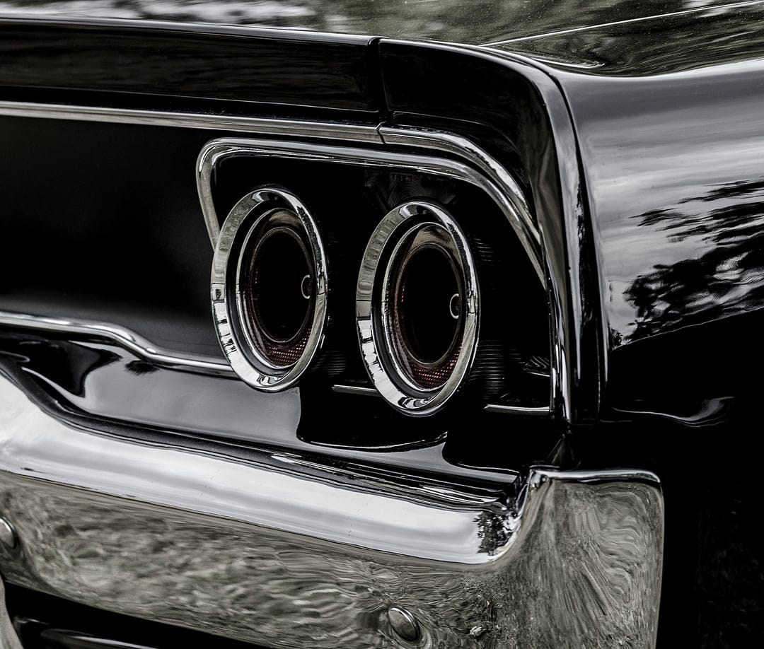 Dual red circular taillights and a polished chrome bumper on a glossy black vintage car in a vertical close-up.