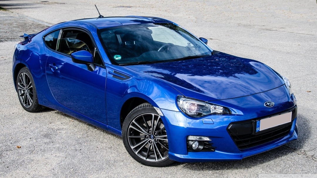 Blue Subaru BRZ sports car parked on grey gravel with metallic reflections, sharp headlights, and alloy wheels.