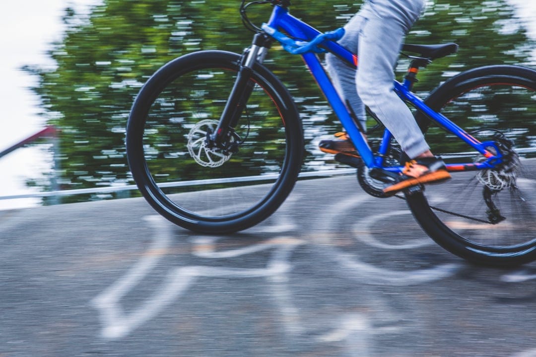 Person on a blue bicycle with orange shoes speeds past blurred trees on a paved path with white chalk markings.