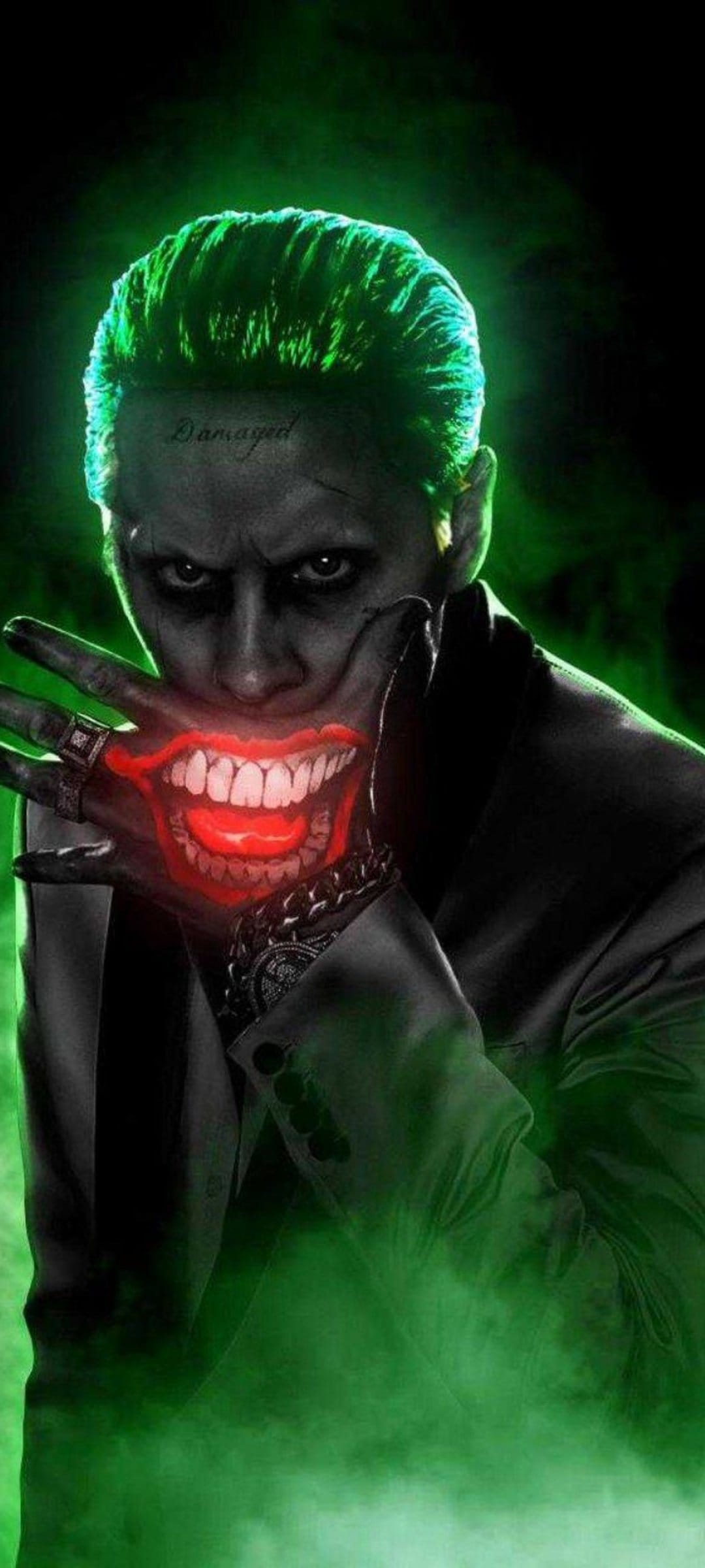 Joker with green hair holds a hand to his face, showing a red smile on his palm and a forehead tattoo.