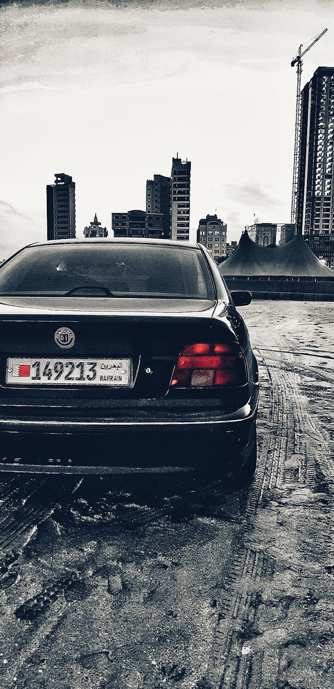 Black BMW sedan rear view on wet asphalt with Bahrain skyscrapers and a construction crane in the background.