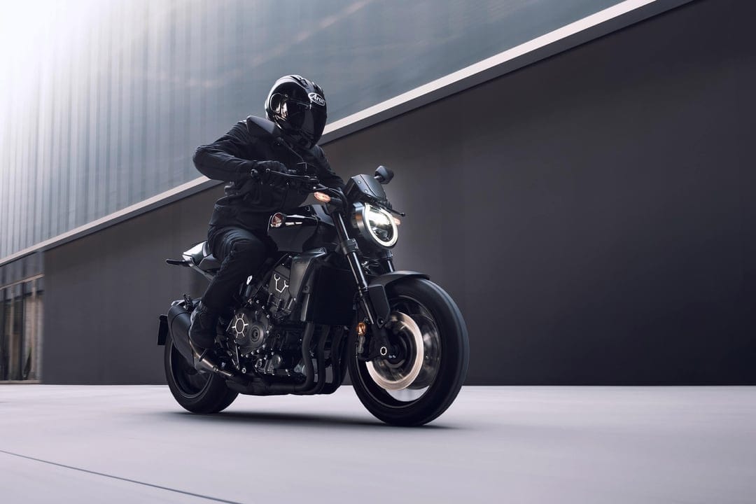 Black naked motorcycle with a rider in matching gear moving past a dark minimalist building at speed.