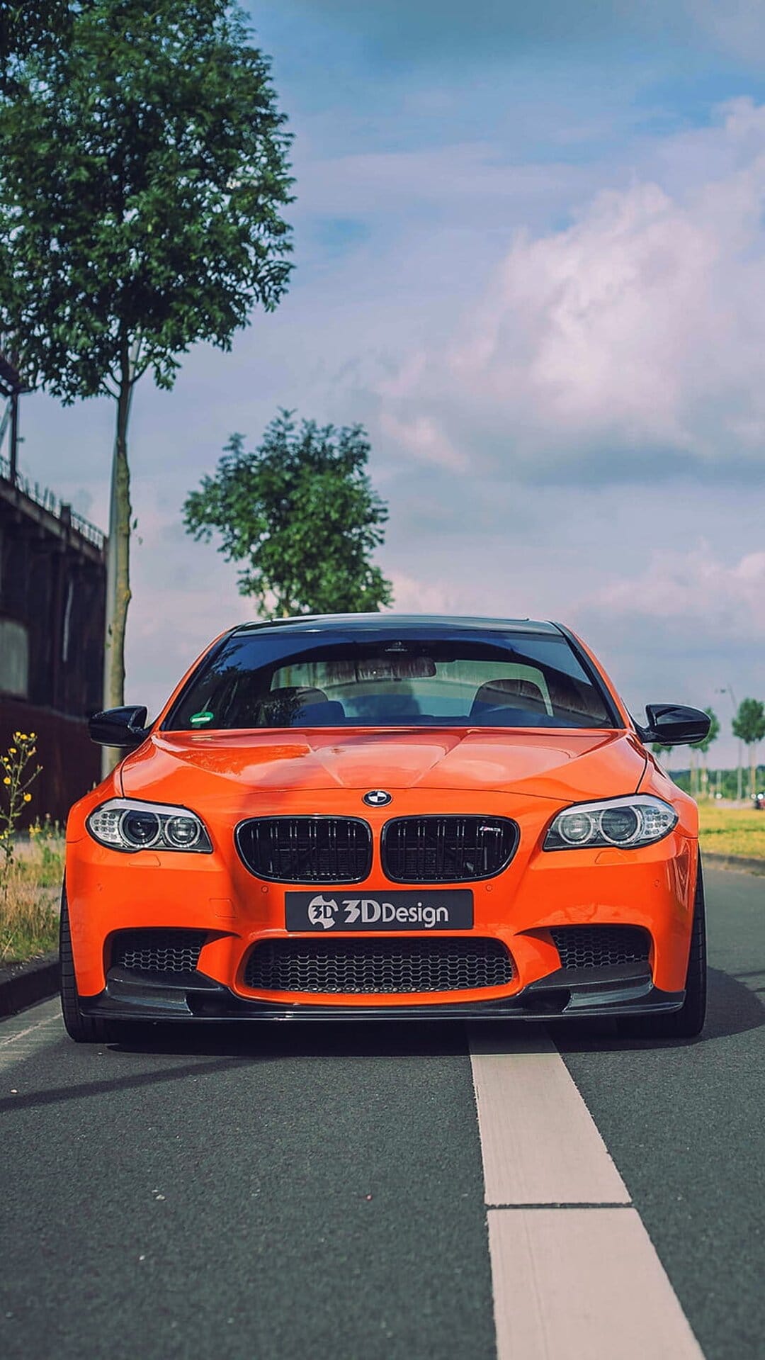 Orange BMW M5 with a 3DDesign body kit parked on a road under a cloudy sky with a bridge in the distance.