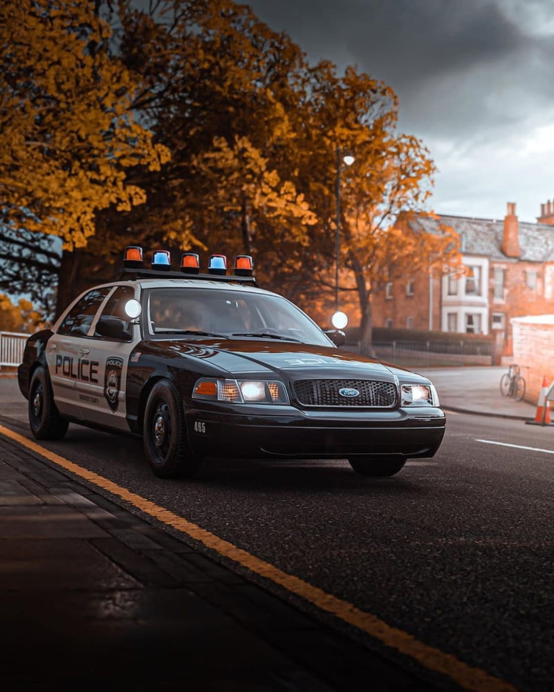 Police car with flashing blue and red lights driving on a wet road lined with orange autumn trees and houses.