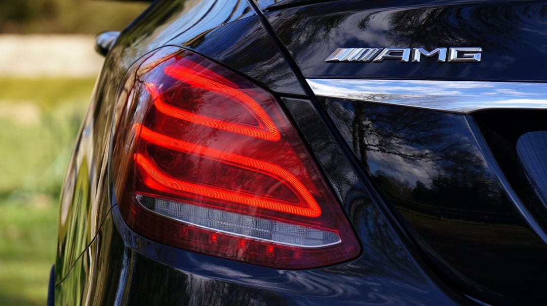 AMG emblem and red LED taillight on a glossy black car reflect green trees in high-resolution detail.