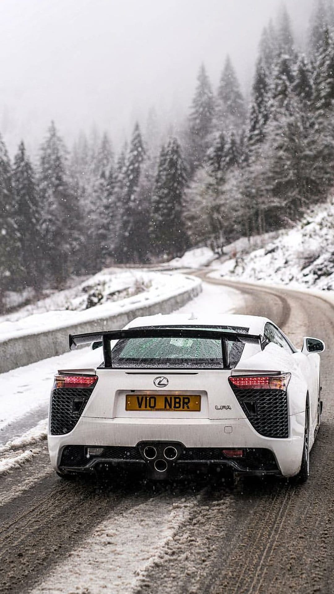 White Lexus LFA supercar rear view driving on a snowy mountain road lined with pine trees during a winter storm.