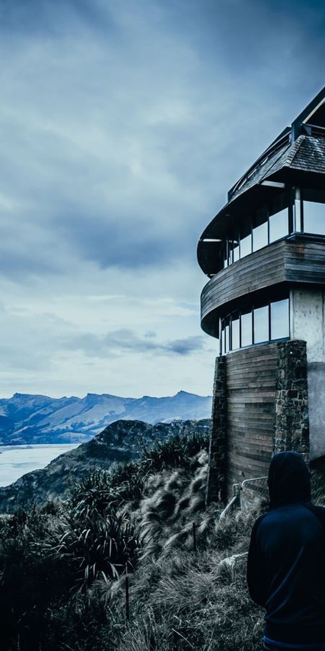 Modern circular building on a rocky cliff overlooking a blue lake and rolling hills under a cloudy sky.