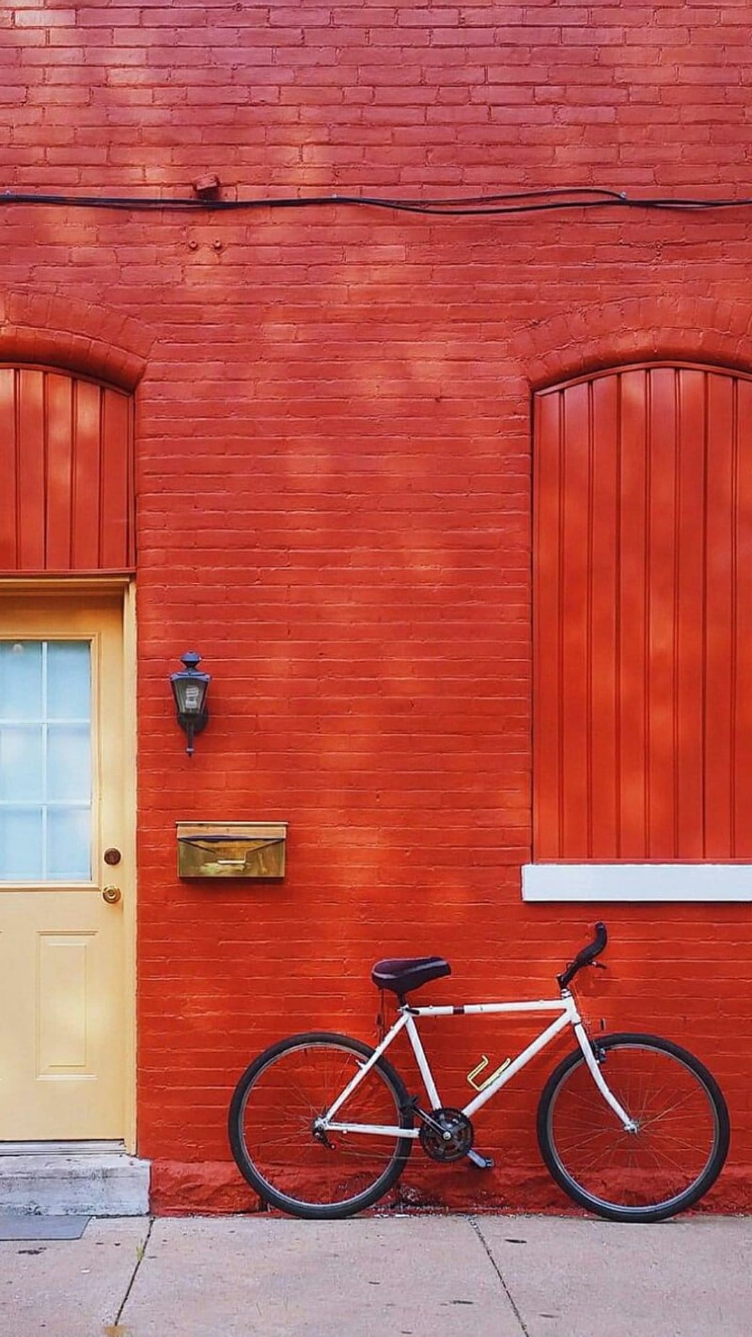 White bicycle leans against a textured red brick wall next to a bright yellow door with a vintage lantern.
