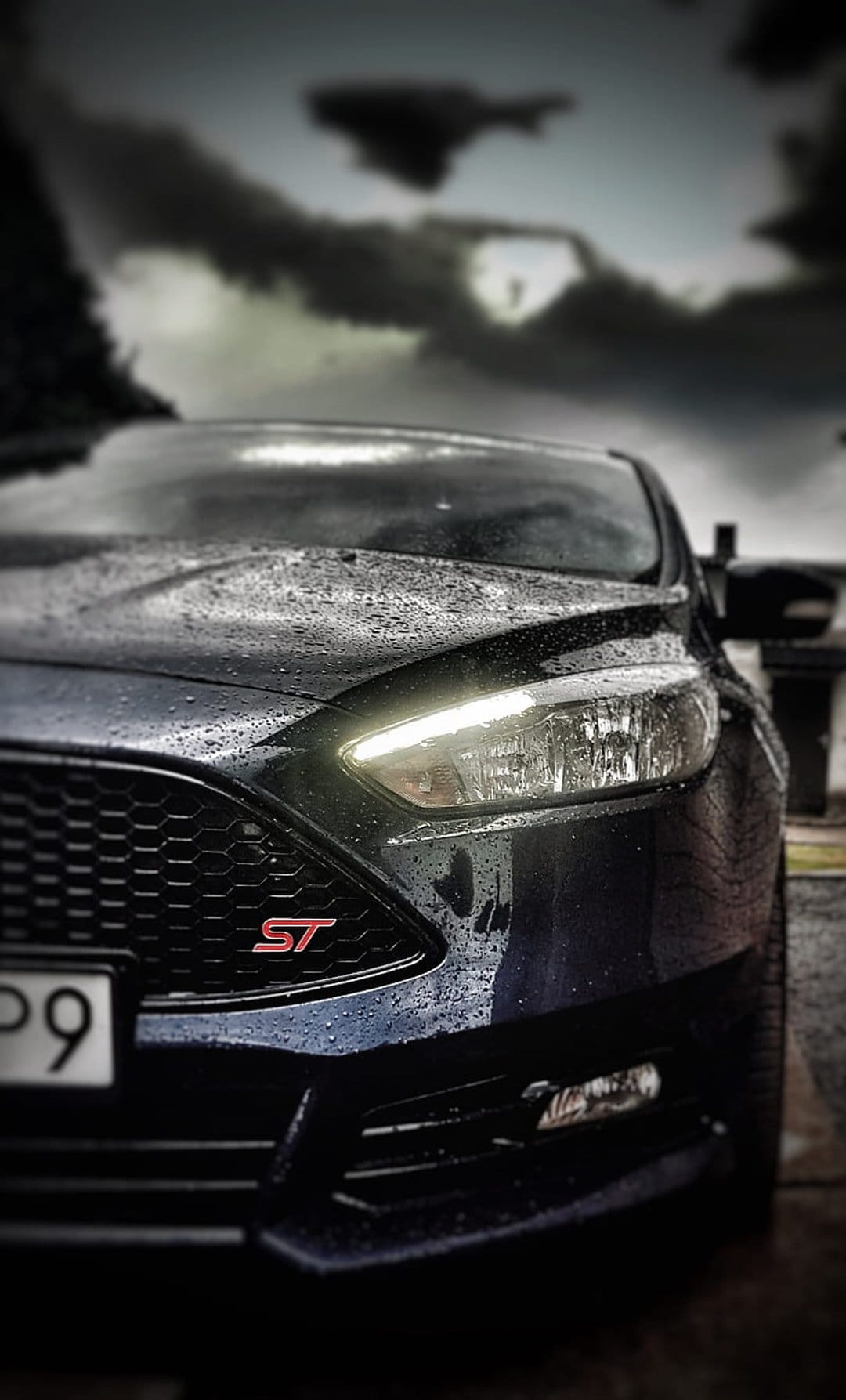 Ford Focus ST headlights glow through rain droplets on a dark blue hood under a stormy sky in a low-angle view.