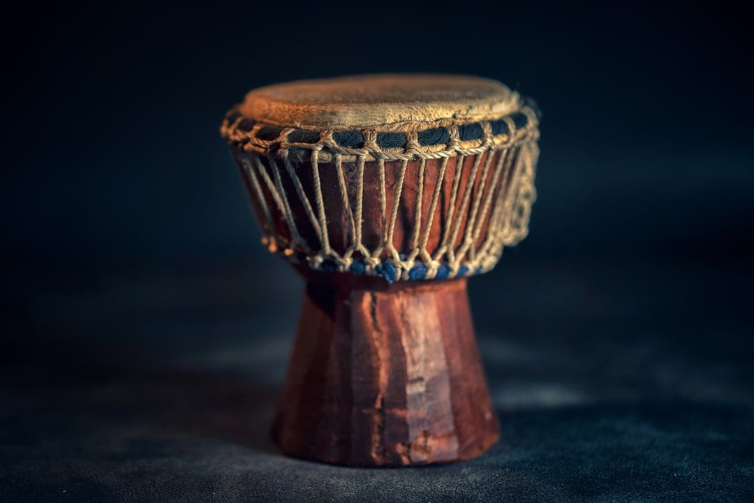 African djembe drum with weathered skin and rope lacing set against a dark, blurred mahogany wood backdrop.