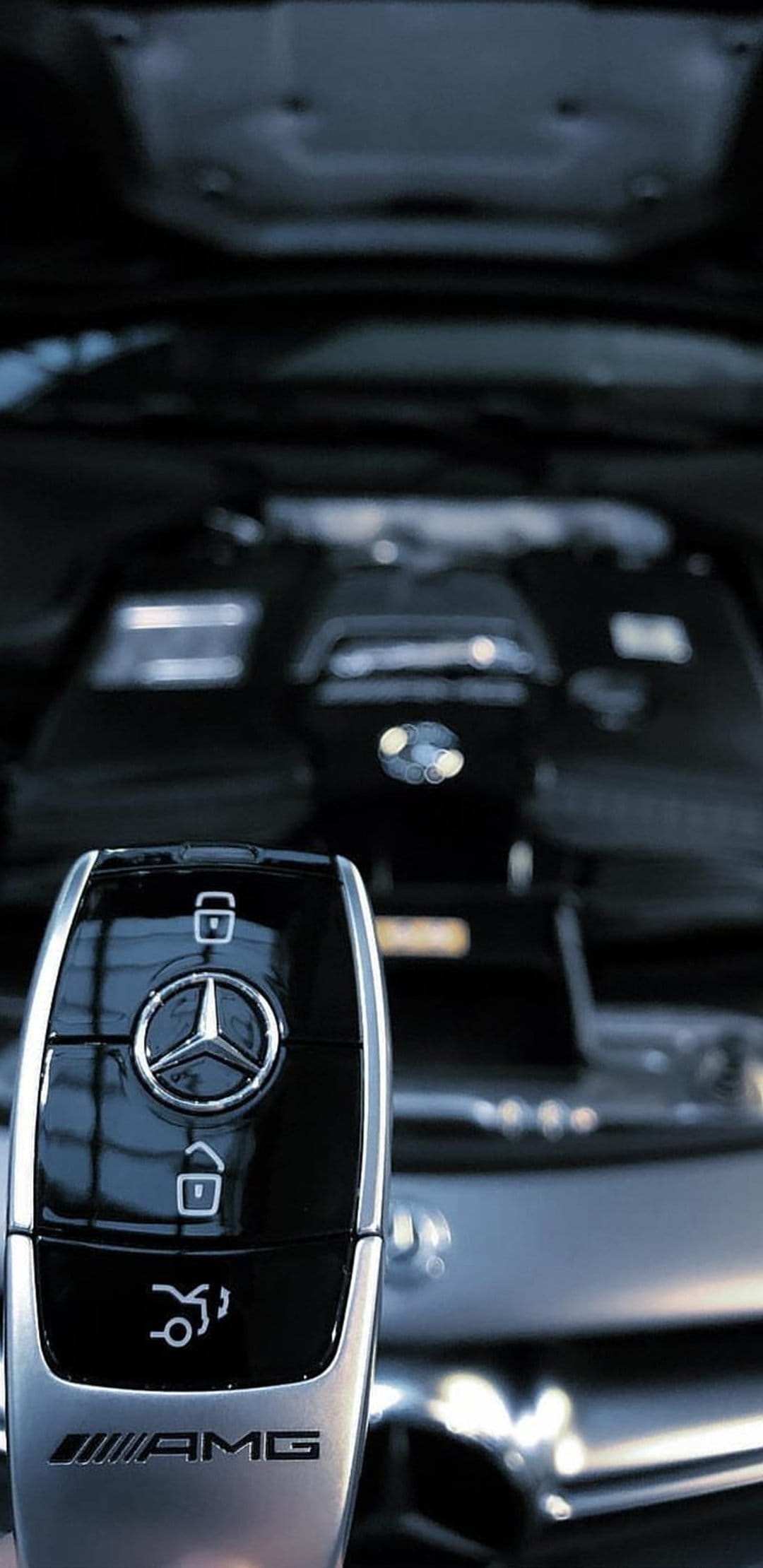 Mercedes-AMG key fob with silver logo centered before a blurred vehicle engine with metallic parts and depth.